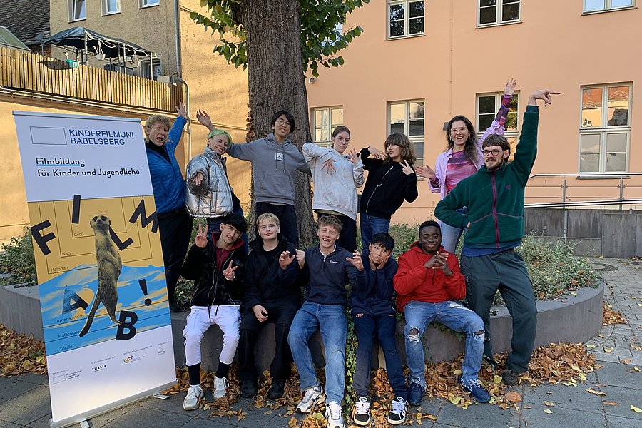 Eine Gruppe Jugendlicher steht vor einem großen Baum für ein Gruppenbild zusammen. Alle posieren zusammen und lächeln. Neben der Gruppe steht ein Rollup das auf den Filmworkshop, den die Jugendlichen gerade gemeinsam besuchen,  hinweist.